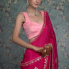 A woman modeling a deep pink silk saree with elaborate gold and silver embellishments, paired with a light pink sleeveless blouse and emerald green and diamond jewelry.