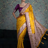 A woman modeling a golden yellow silk saree with intricate gold and purple borders and a purple blouse.